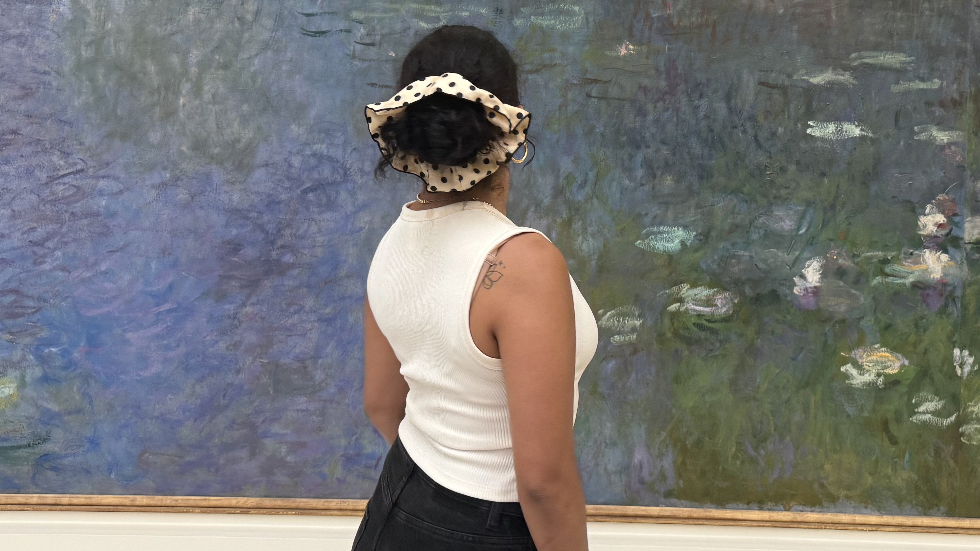 Woman wearing a white sleeveless top and dark pants stands in front of an impressionist water lily painting, hair tied with a large polka-dot scrunchie.