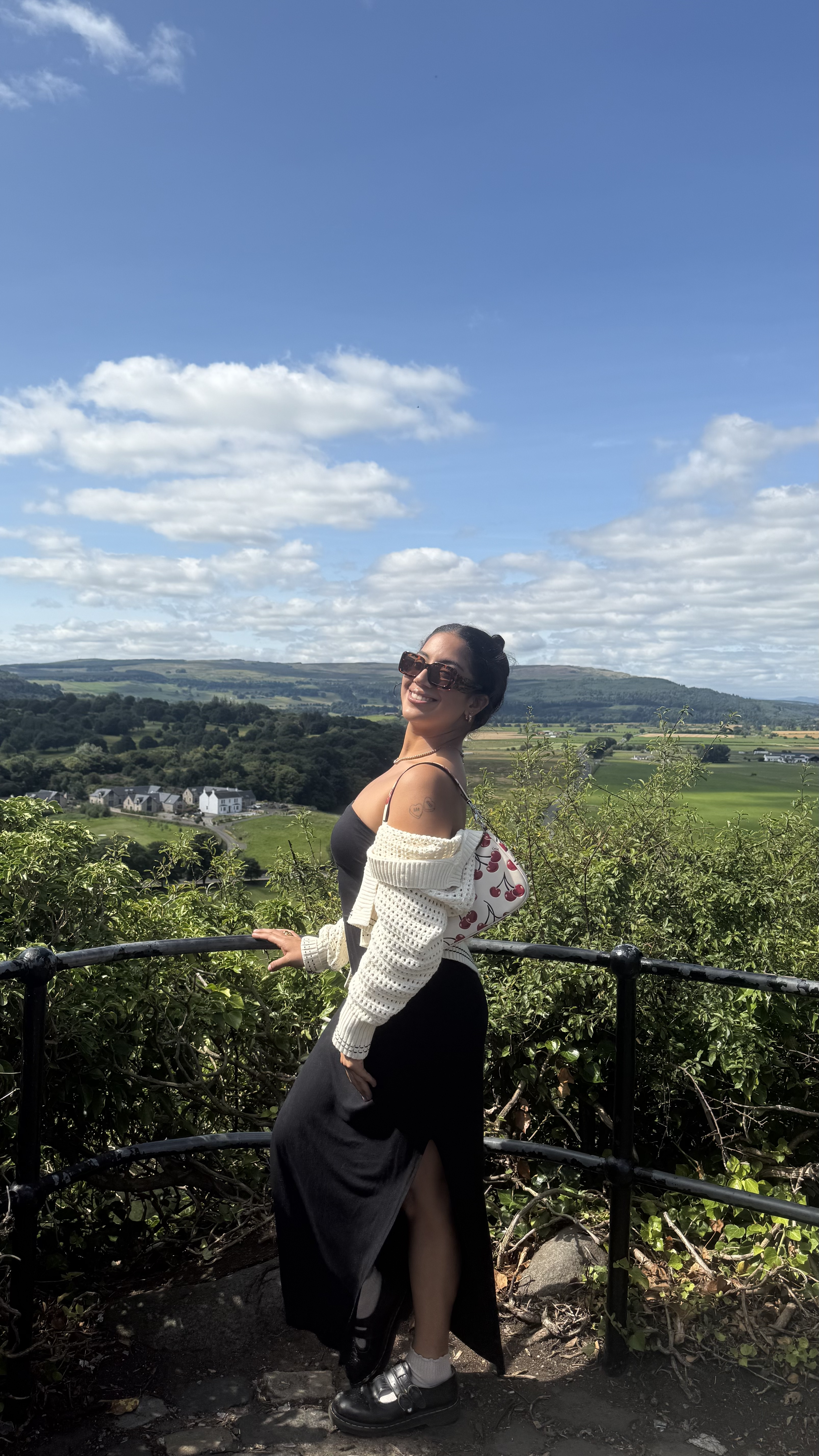 A woman standing outdoors on a scenic overlook with lush greenery and rolling hills in the background under a bright blue sky with scattered clouds. The woman is wearing a black dress with a high slit, a light knit cardigan draped over the shoulders, and sandals, holding a cherry patterned purse.