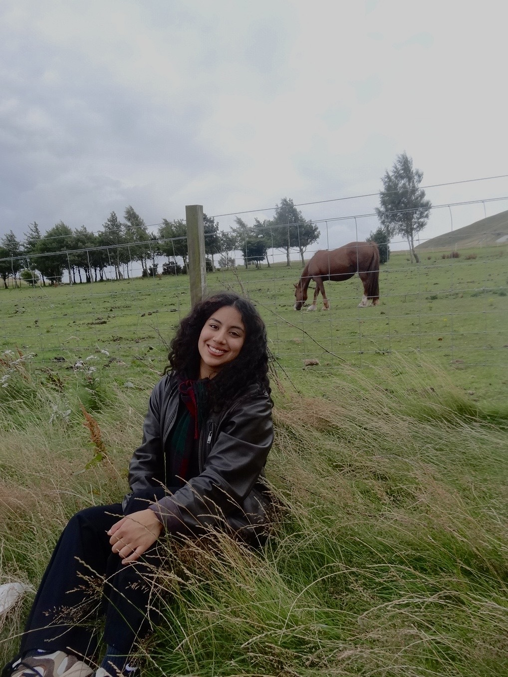 A woman is sitting on tall grass near a wire fence in a green pasture. She is wearing a dark leather jacket and dark pants, with hands resting on her knees. In the background, a brown horse is in the field, and there are trees along the horizon under a cloudy sky.